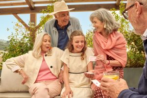A casual outdoor gathering featuring four people sitting together on a sofa. A woman holds a playing card while smiling at a teenage girl. An older man and woman look on, with greenery and a pergola in the background, capturing a lively moment at Casa Barbara.