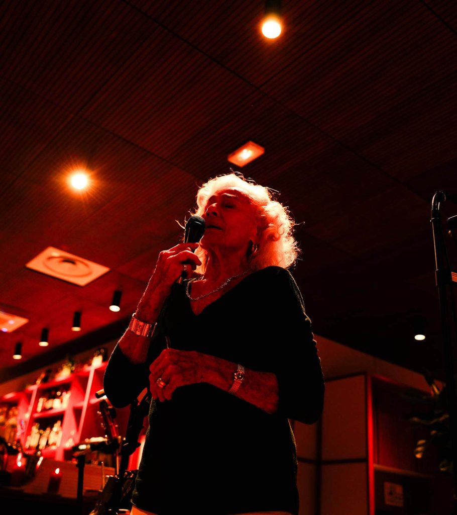 Une femme âgée chante au micro sur une scène, entourée de lumières orangées, ambiance de spectacle ou soirée musicale dans un espace cosy. Une femme âgée chante au micro sur une scène, entourée de lumières orangées, ambiance de spectacle ou soirée musicale dans un espace cosy.